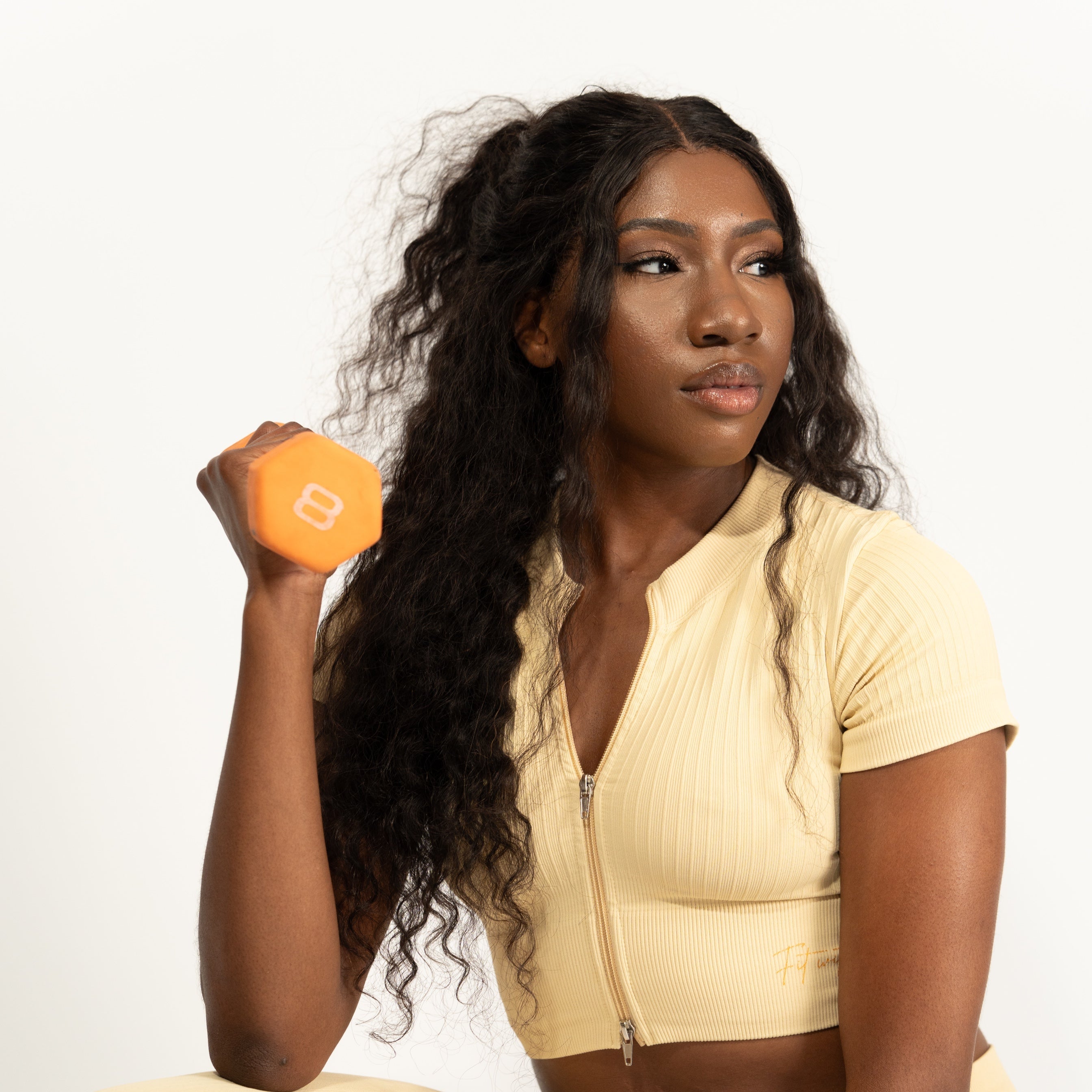 A woman crouching in a matching pale yellow ribbed activewear set, featuring a short-sleeve zip-front crop top and leggings, while holding a small orange hexagonal dumbbell against a plain white studio background.